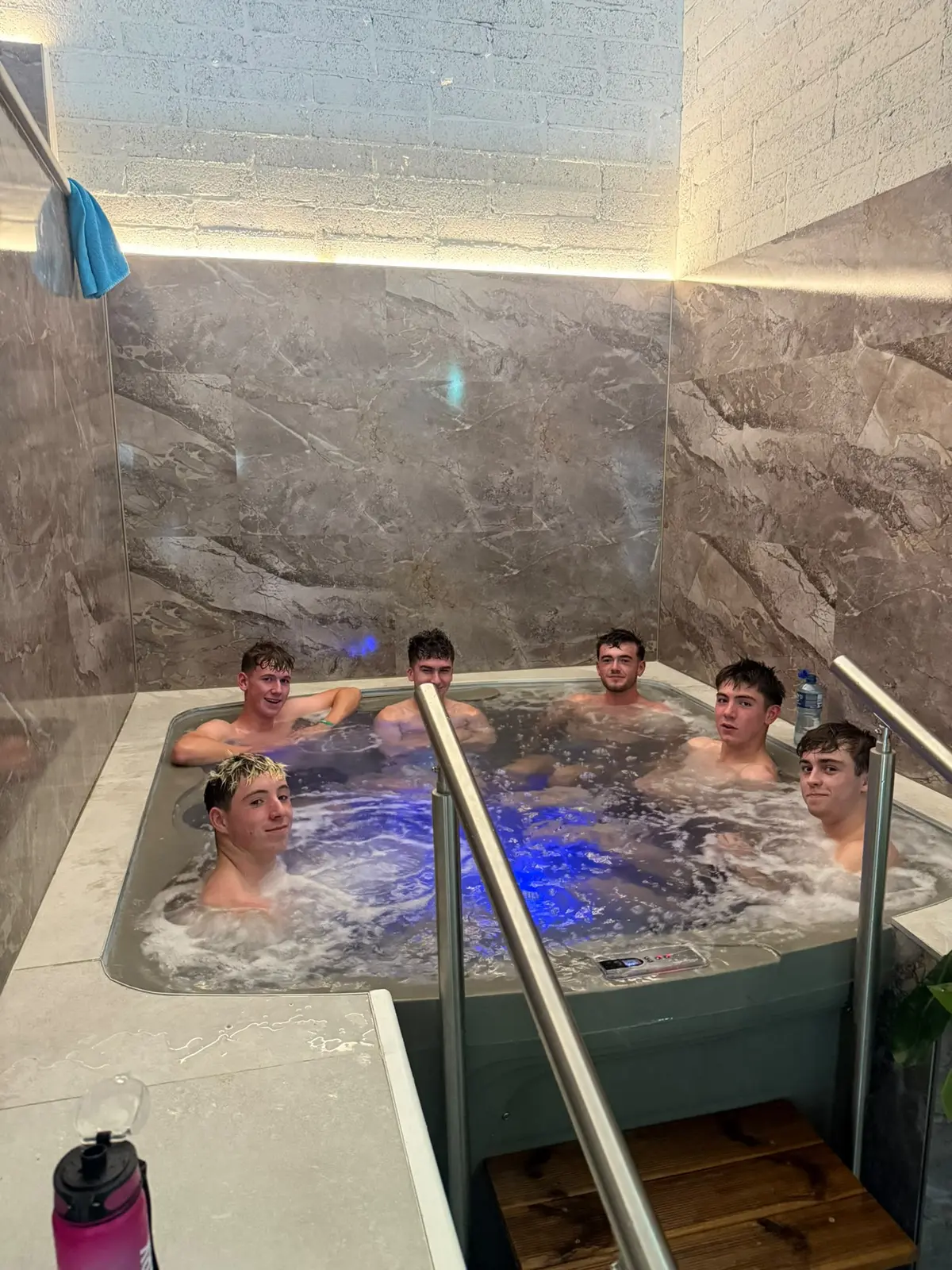 A group of five in the jacuzzi, the water lit blue, towels on the rail