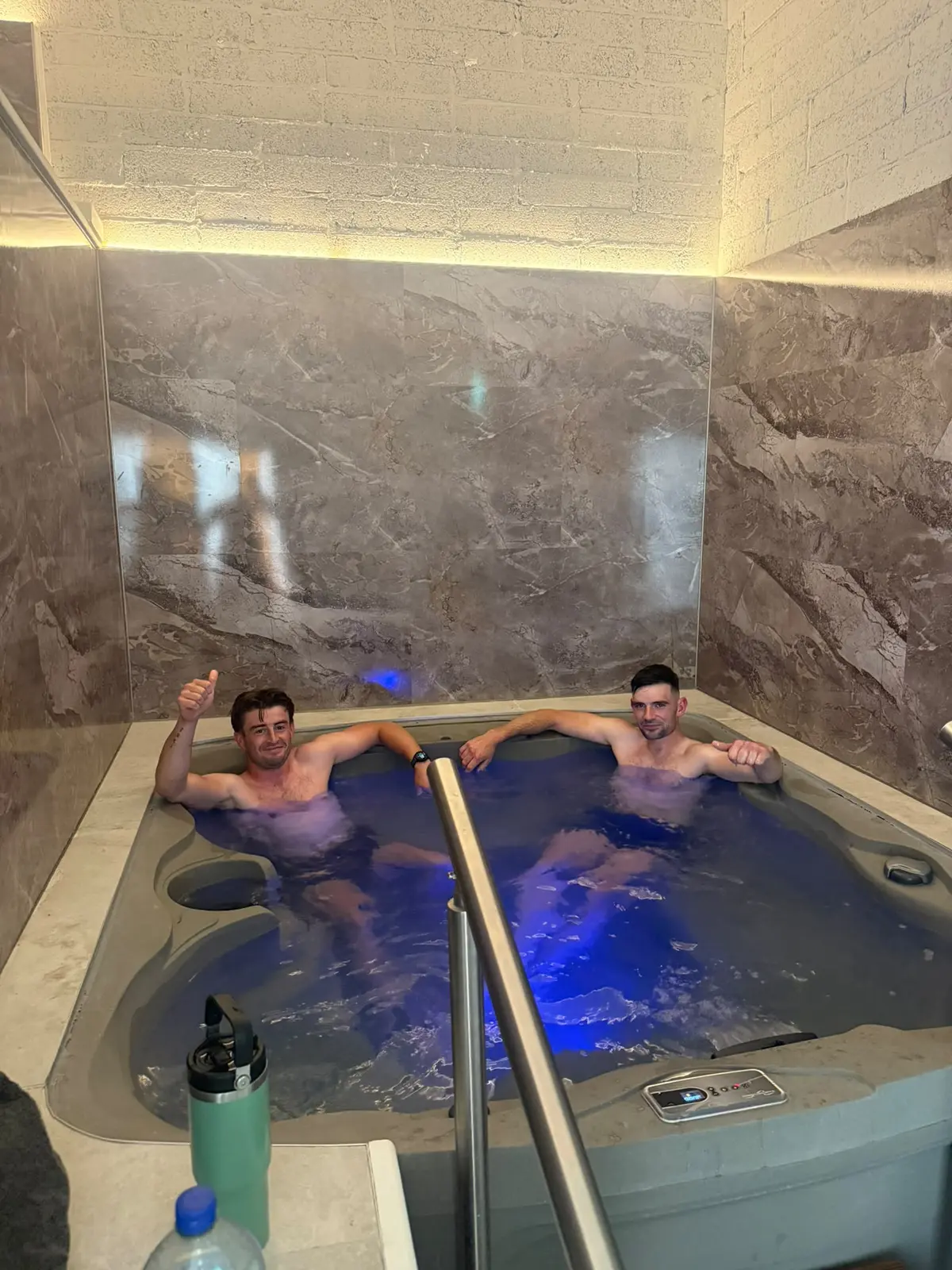 Two guests in the jacuzzi with the blue light on, a water bottle on the floor beside the steps