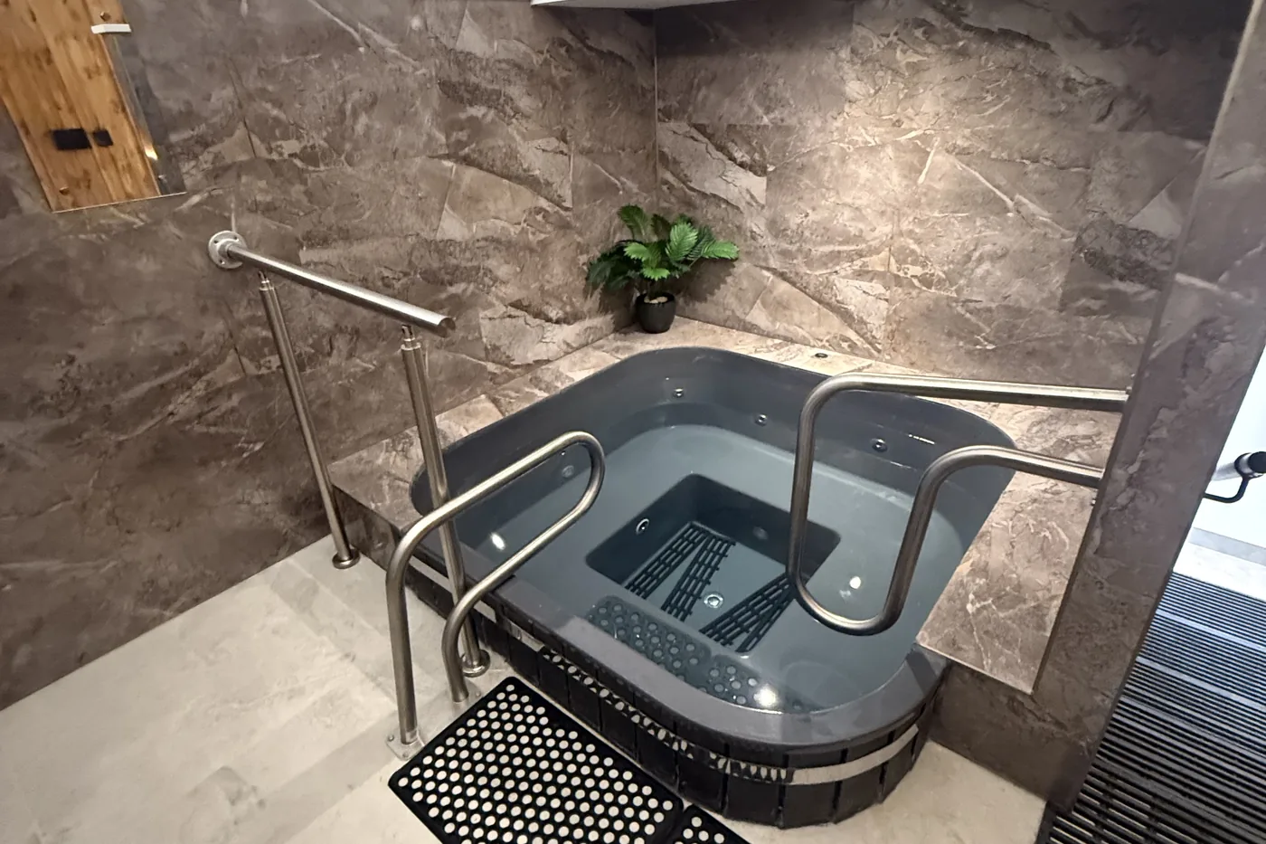 The cold plunge: a grey tub set into grey marble tiles, stainless steel handrails and black rubber mats on a pale stone floor