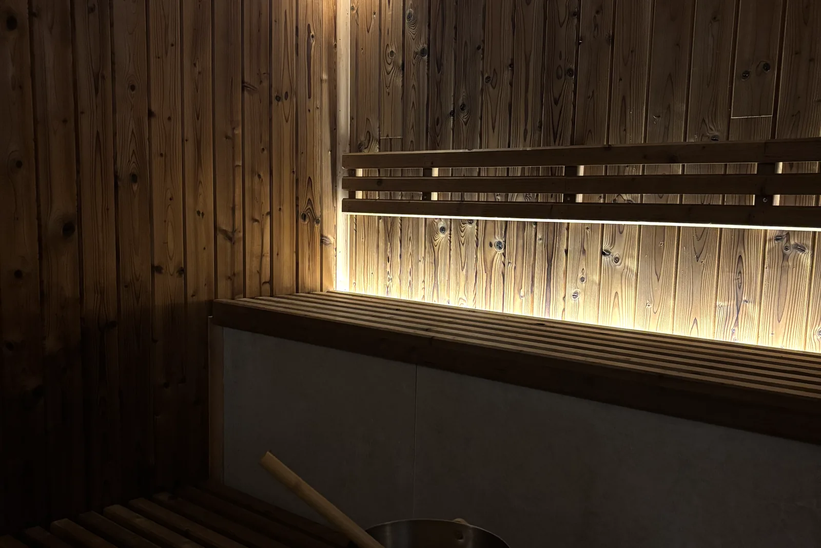 Inside the sauna: slatted wooden benches beneath a backlit wooden rail, a wall lamp and the water bucket